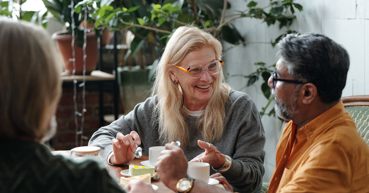 Three older adults seated at a table in a bright indoor setting, sharing coffee and dessert while engaged in conversation, with leafy houseplants and natural light in the background.